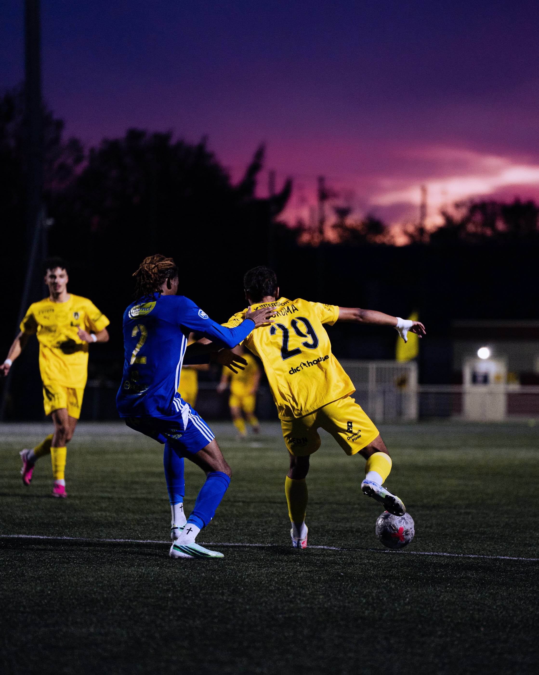 Un joueur de l'ASPTT Caen, maillot jaune n° 29, dribble un adversaire sous un ciel de coucher de soleil, à la Hache